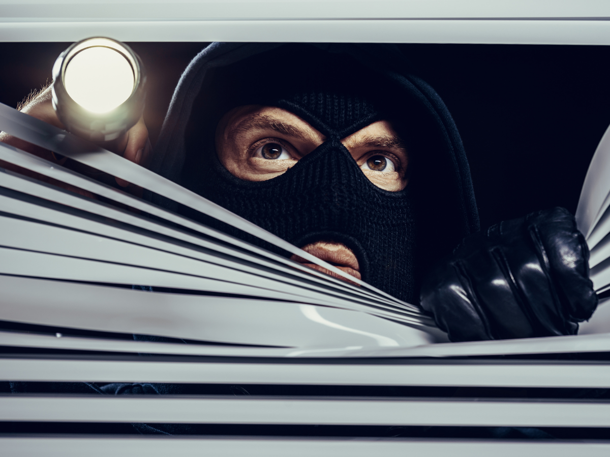 Burglar wearing a black mask and gloves peeking through blinds in a window with a flashlight.