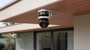 A white and black reolink PTZ security camera mounted on the underside of a wooden roof overhang outside a modern building.