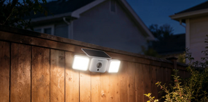 A solar-powered outdoor Reolink camera with floodlight mounted on a wooden fence at dusk, casting a warm glow on the fence while illuminating two bright LED light panels and a small rectangular solar panel above them.