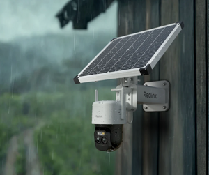 A Reolink solar-powered outdoor security camera mounted on a dark wooden wall, with a tilted solar panel above it. Rain is visibly falling in the background, showing the camera's weather-resistant design.