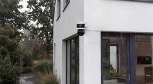 A white and black security camera mounted on the corner of a modern white stucco house, shown outdoors in the rain with trees and a garden visible on the left.