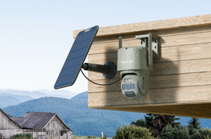 A solar-powered outdoor security camera mounted on a wooden wall, with a small solar panel attached to the same bracket. The camera has a camouflage-style finish and is shown against a rural background with mountains, a barn, and trees.