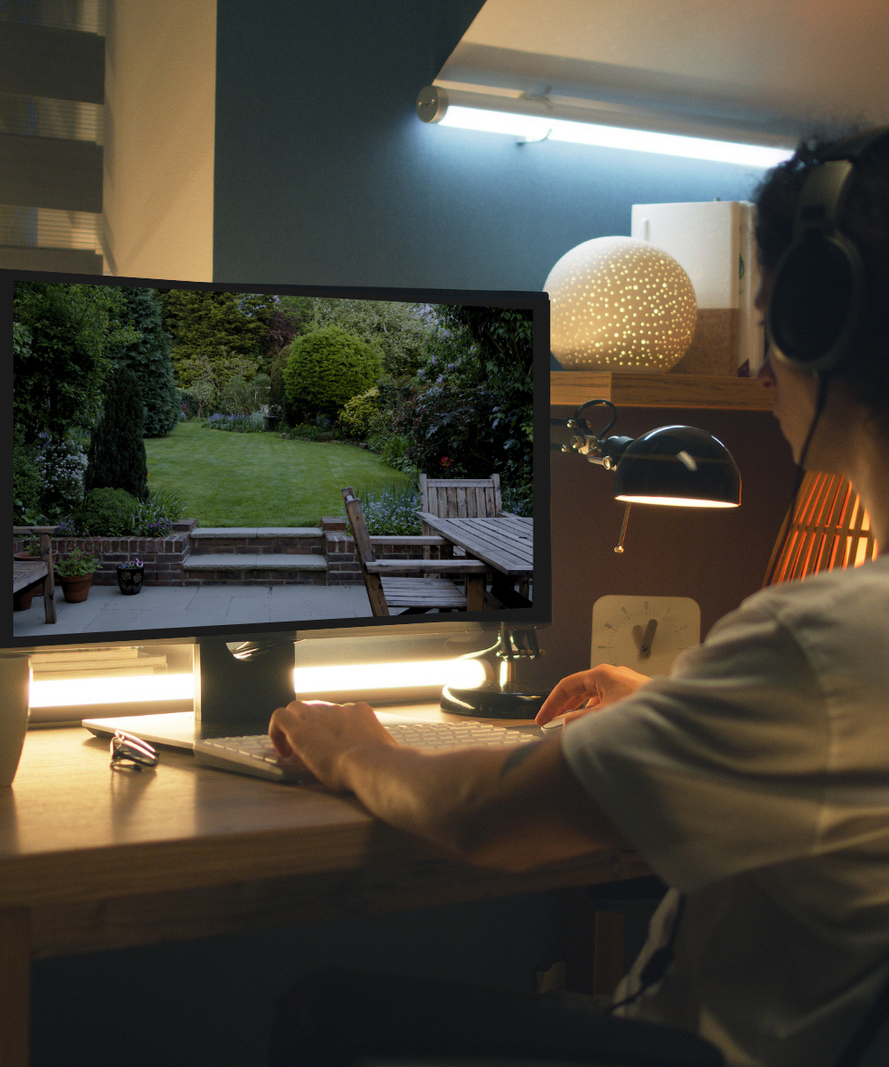 Person sitting at a desk with headphones on, using a computer with a garden cctv view on the screen.